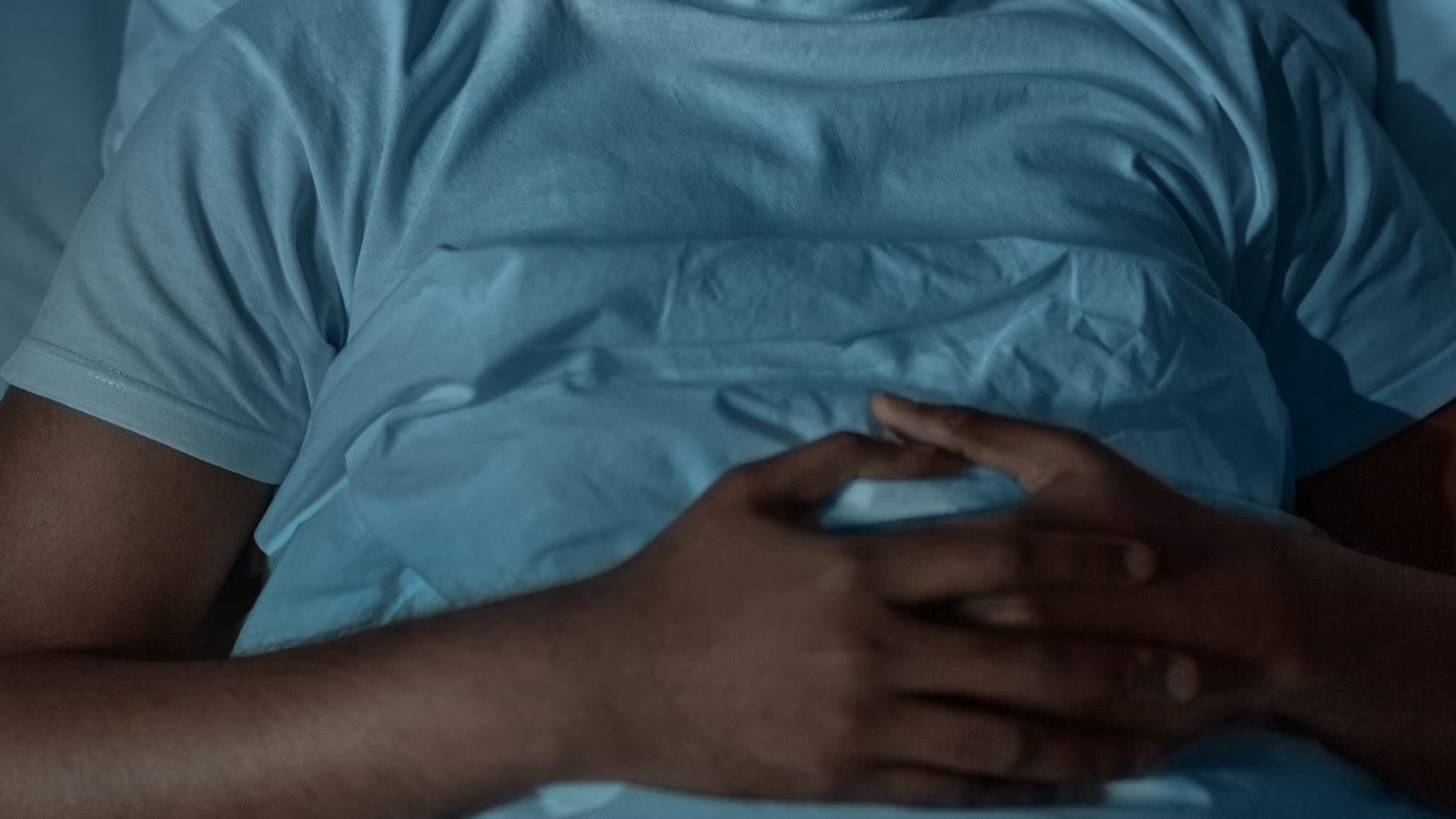 A person lies in bed with hands clasped over a blanket.