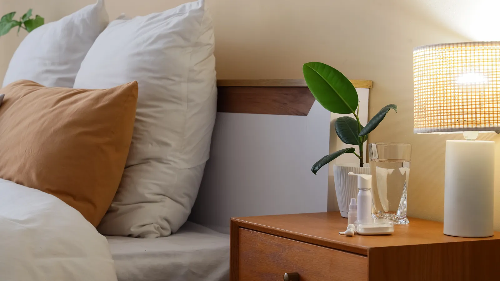 A bedside table with a lamp, a glass of water, a candle and a plant beside a bed