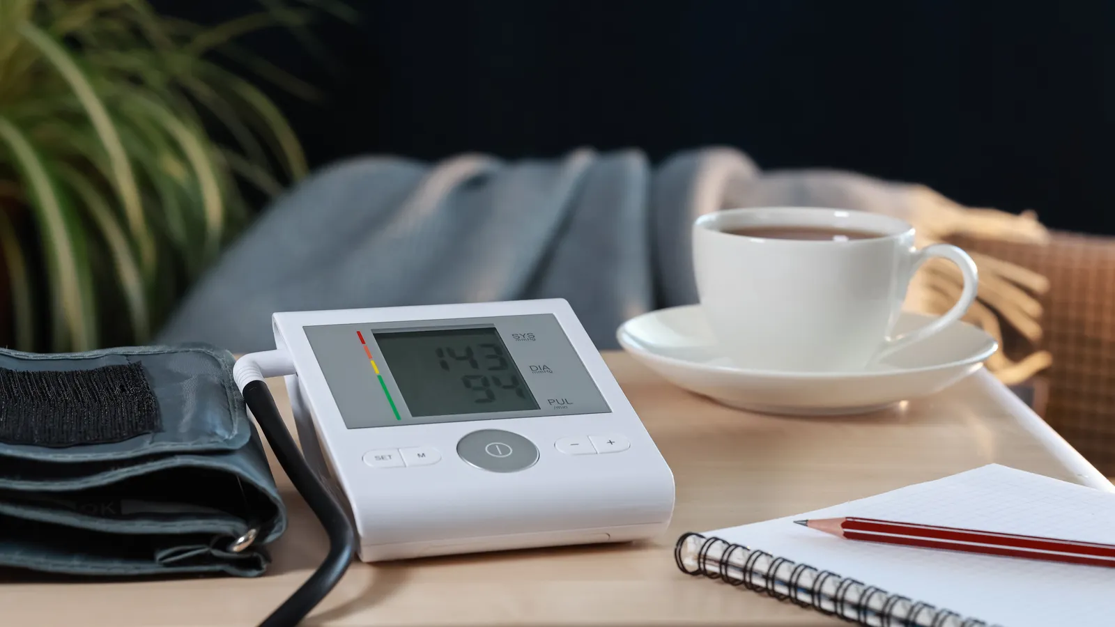 A digital blood pressure monitor with its cuff beside a notebook and a cup of tea on a table