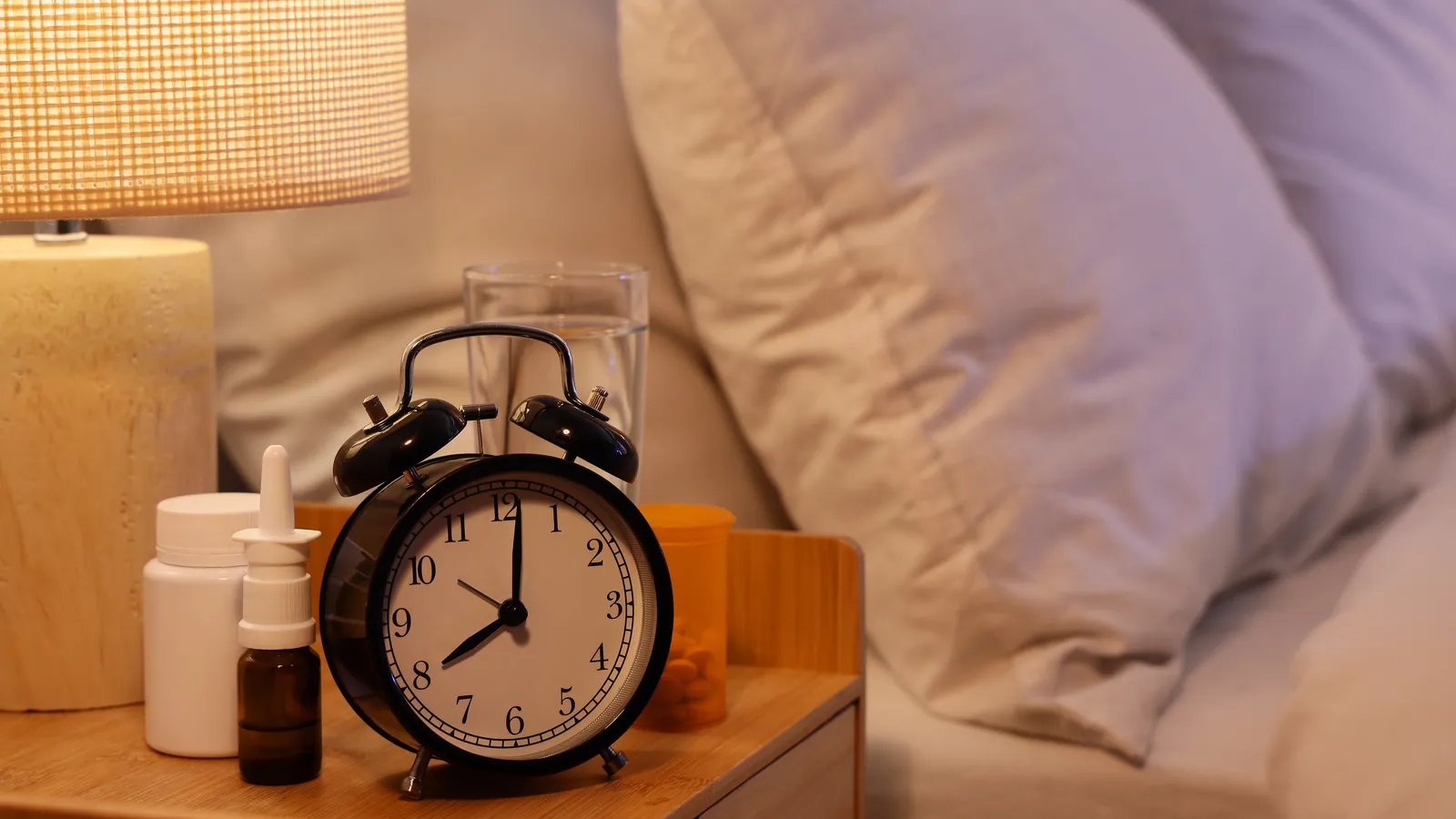 An alarm clock and medicine bottles on a bedside table beside a pillow in warm lamplight