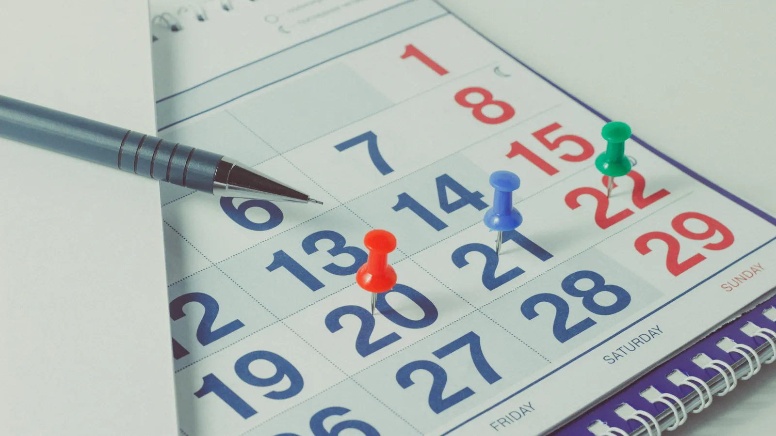 A wall calendar with coloured push pins marking several dates and a pen laid across it