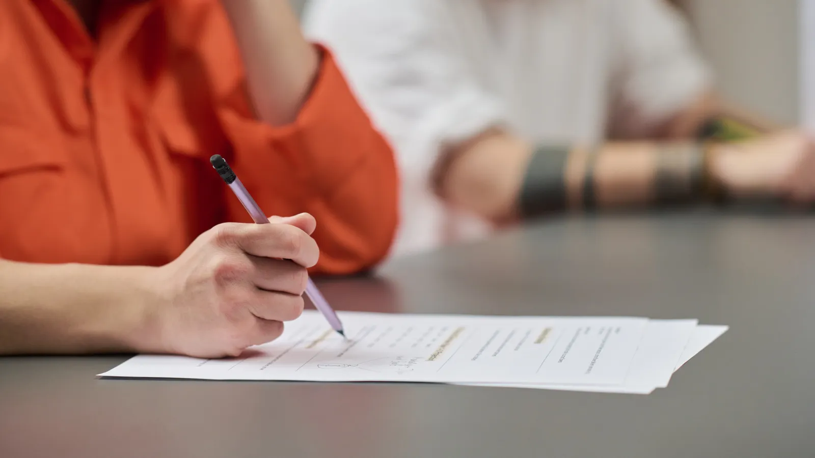 A hand in an orange sleeve writing with a pen on a worksheet at a table