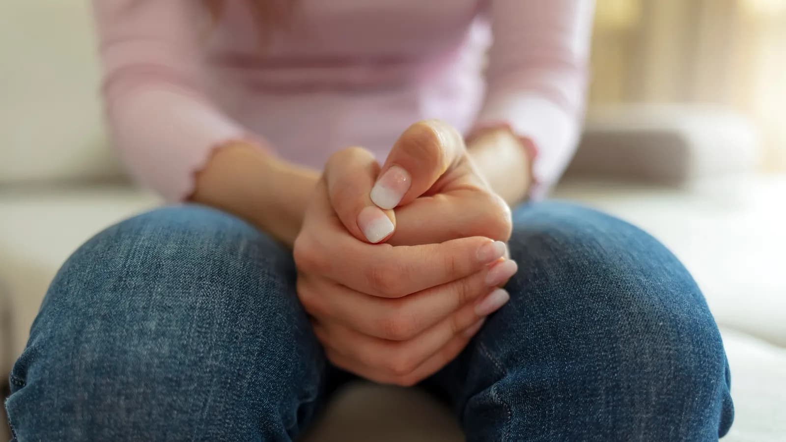 A person's hands clasped tightly in their lap while seated on a sofa