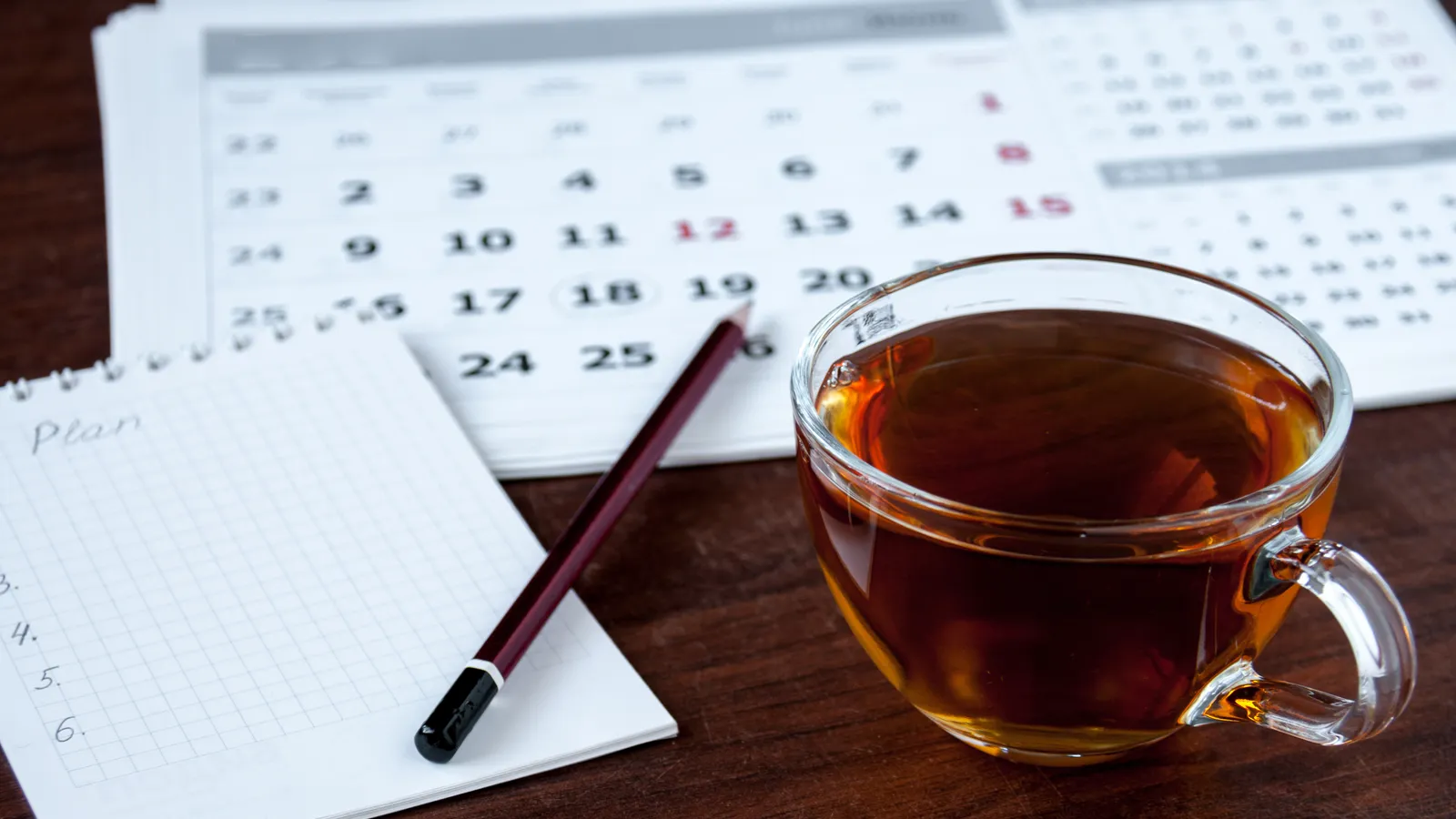 A cup of tea beside a desk calendar and a notepad with a pencil
