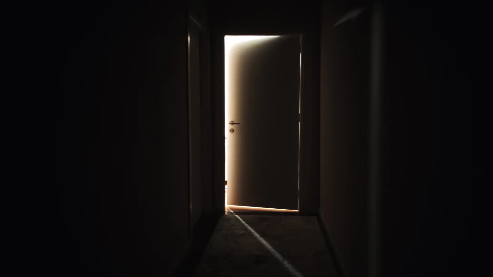 A door standing slightly open at the end of a dark corridor with warm light behind it