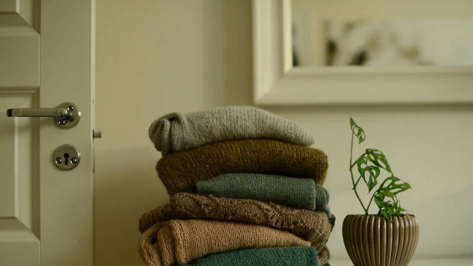 A stack of folded knitted sweaters and a small plant on a white dresser