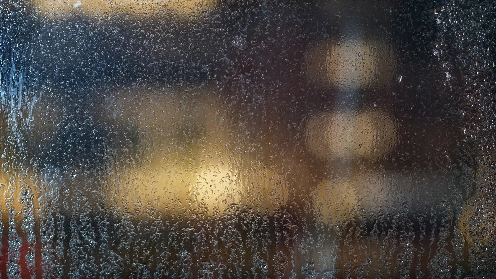 Condensation and rain drops on a window pane blurring the lights behind it