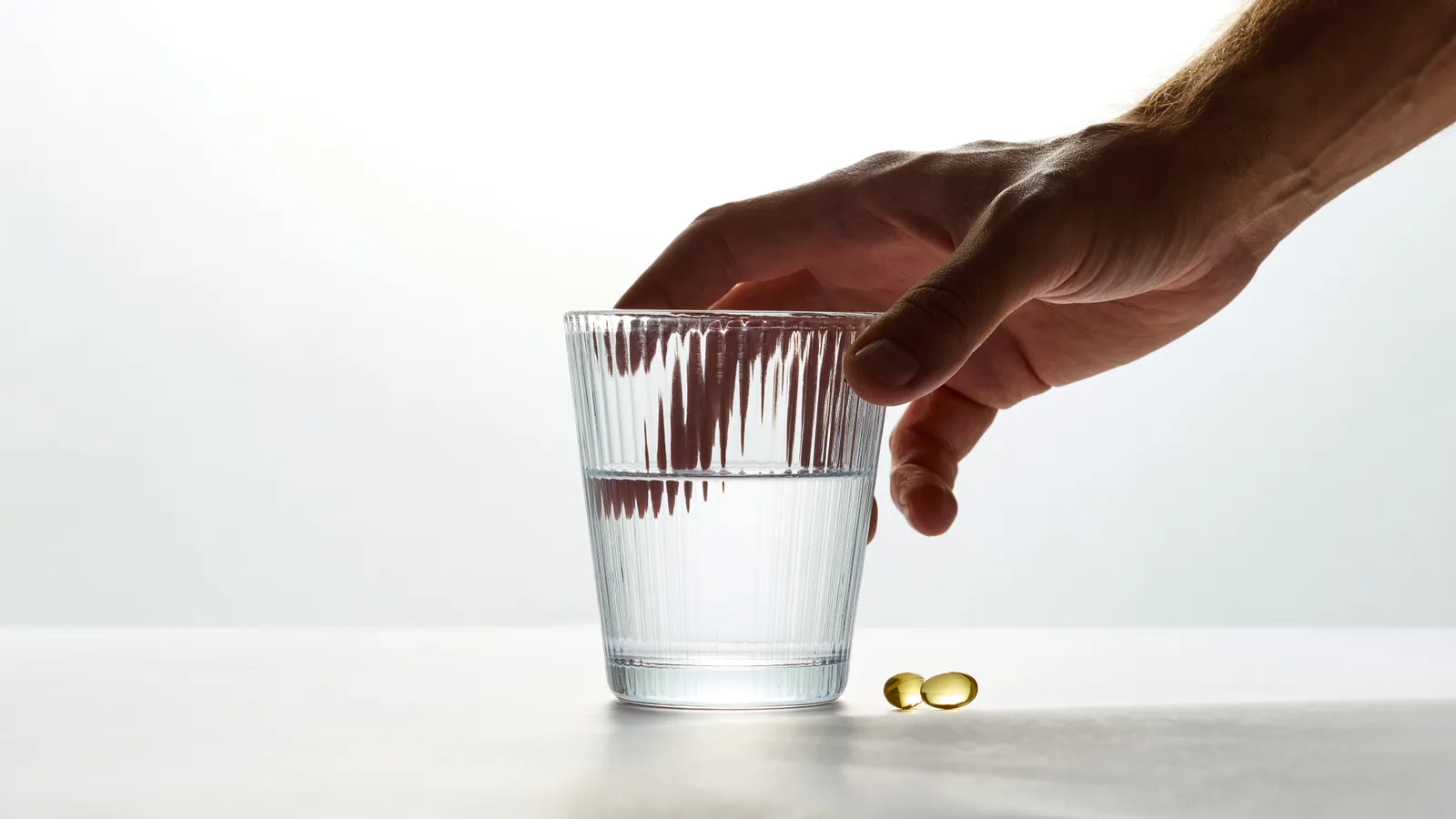 A hand reaching for a glass of water on a white table, with two softgel capsules beside it
