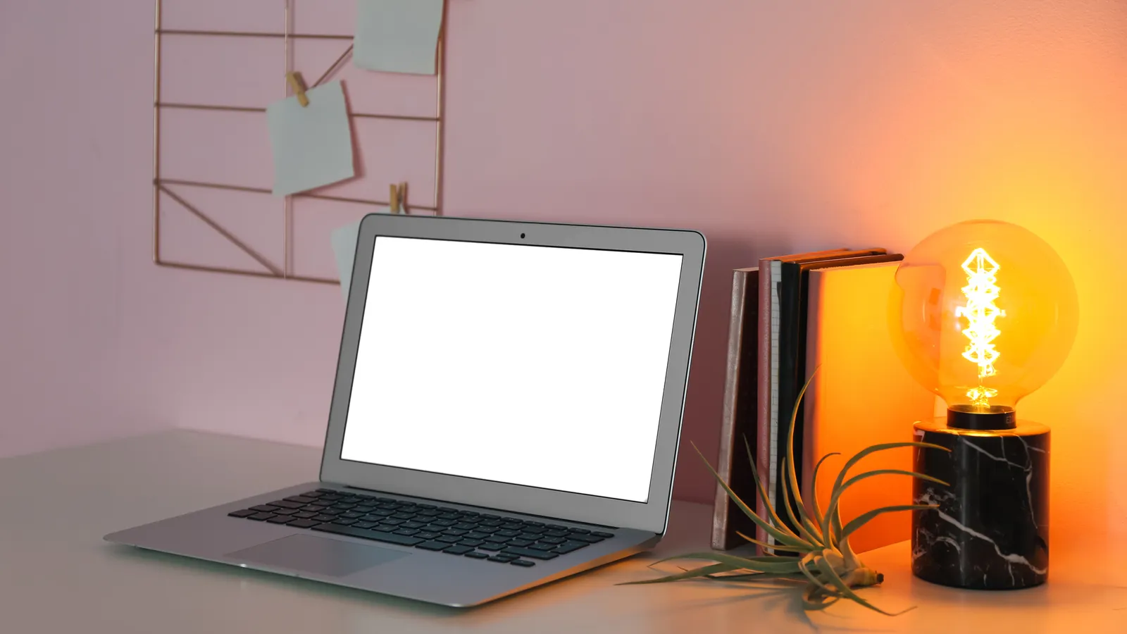 A laptop with a glowing screen beside a lit bulb lamp on a desk at night, with notes pinned on the wall