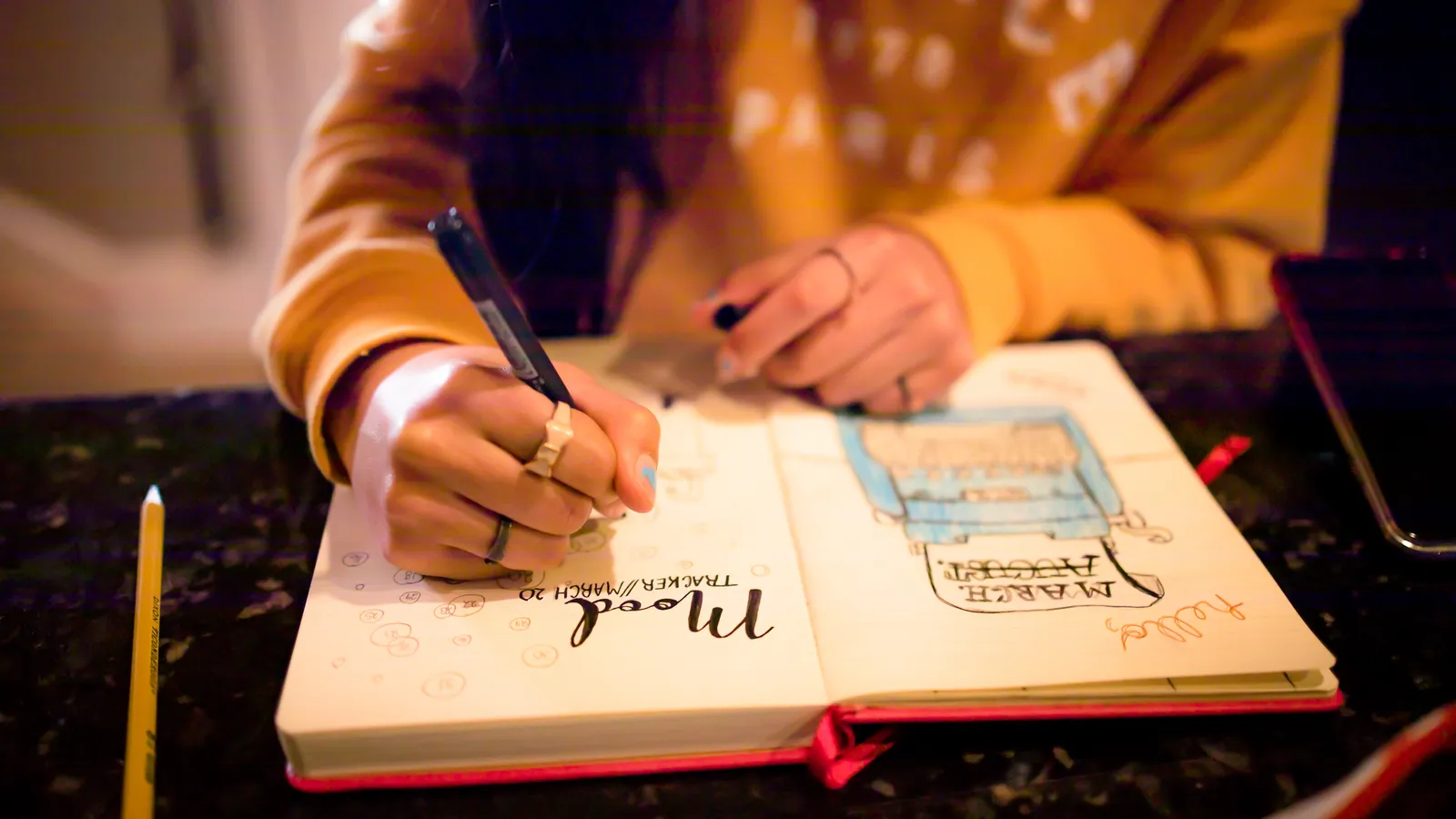 A hand writing with a pen in a bullet-journal mood tracker beside string lights