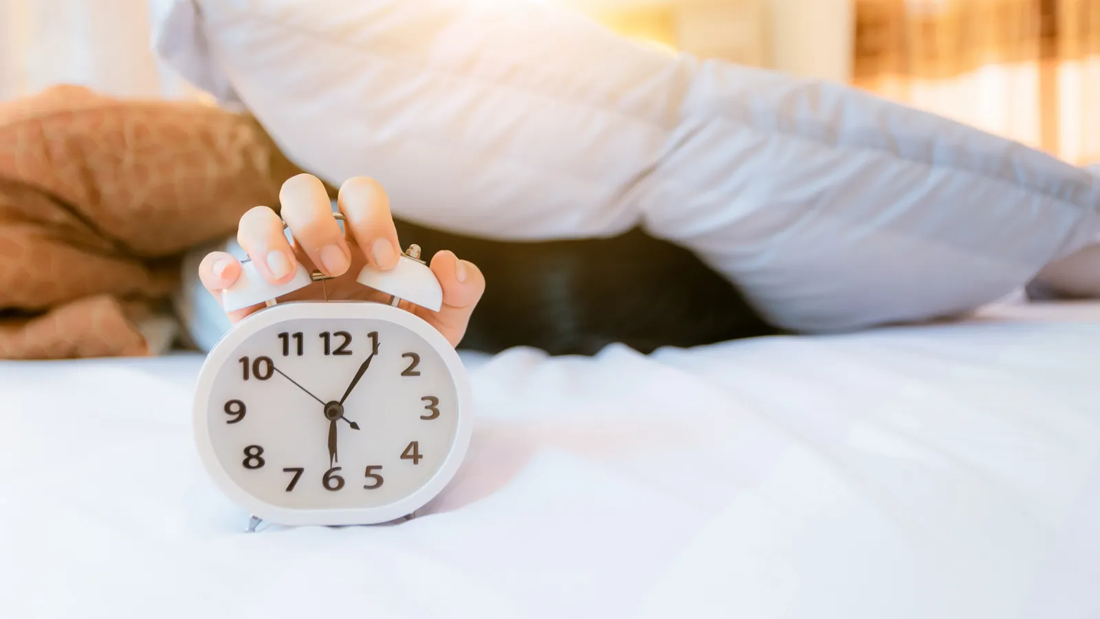 A hand reaching out from under a duvet to an alarm clock on the bed in morning light