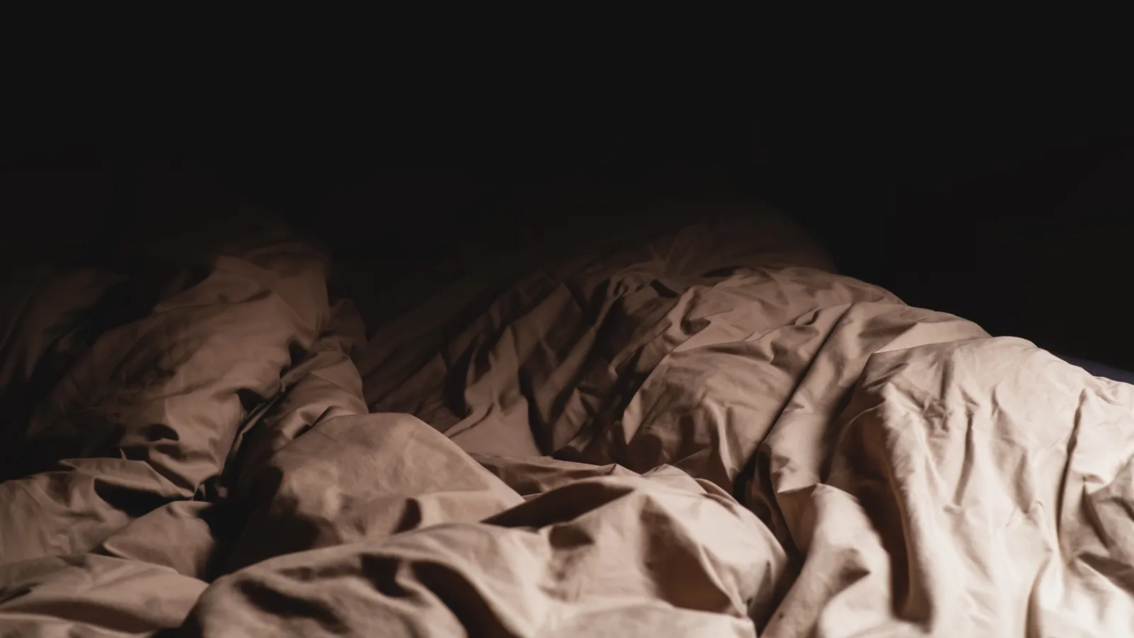 An empty unmade bed with a crumpled blanket in a dark room lit by a lamp