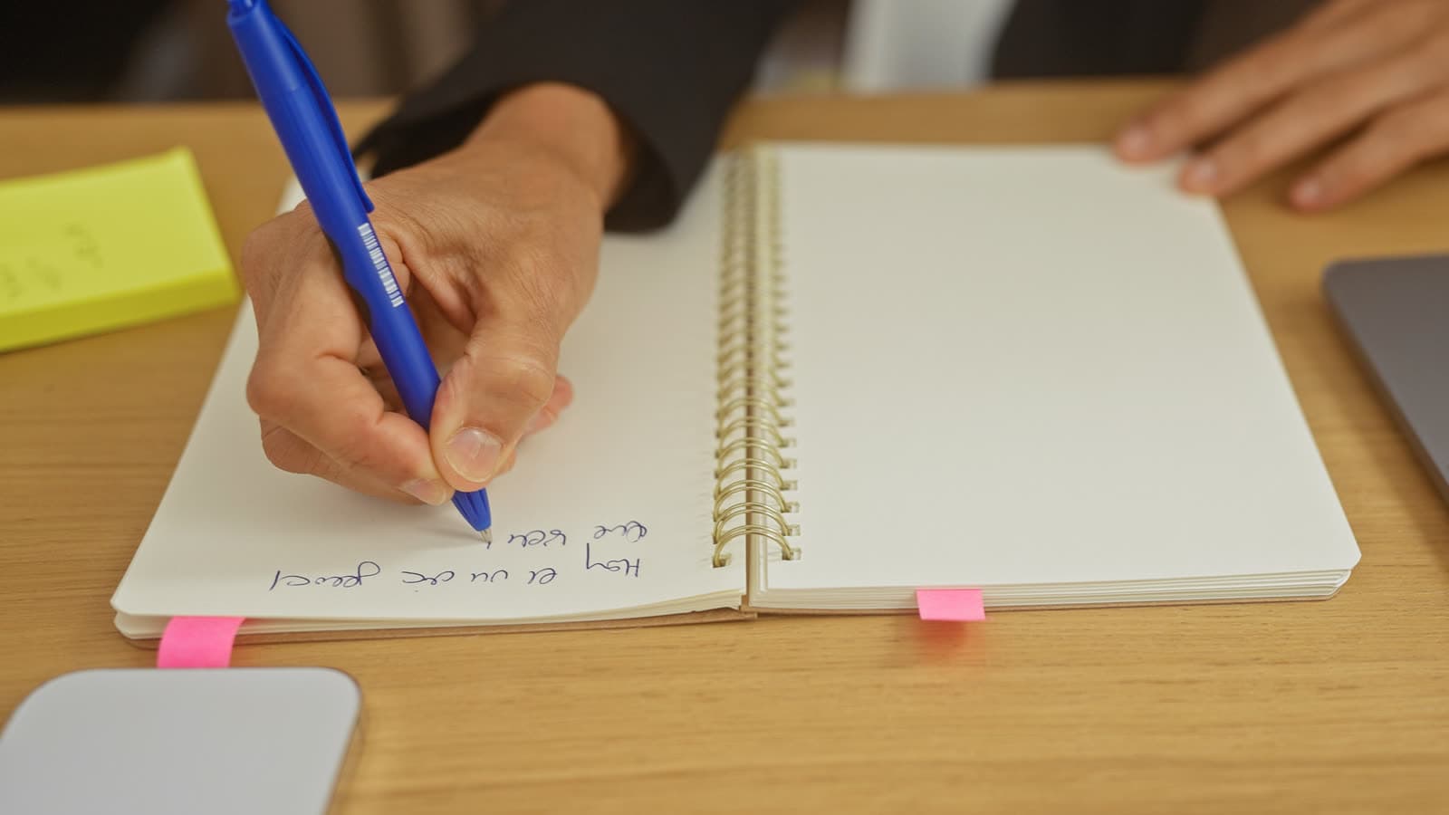 A hand writes in a notebook beside a laptop.