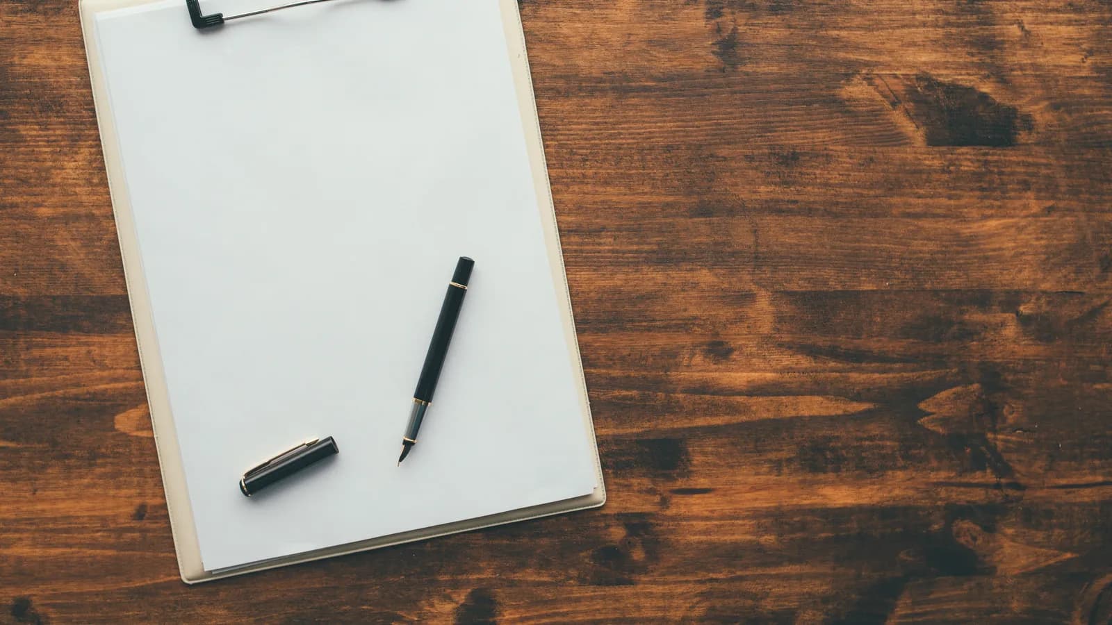 A blank clipboard with a sheet of paper and a pen on a dark wooden table