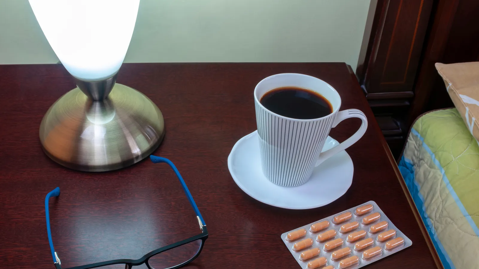 A bedside lamp, a cup of coffee, glasses and a blister pack of tablets on a wooden nightstand