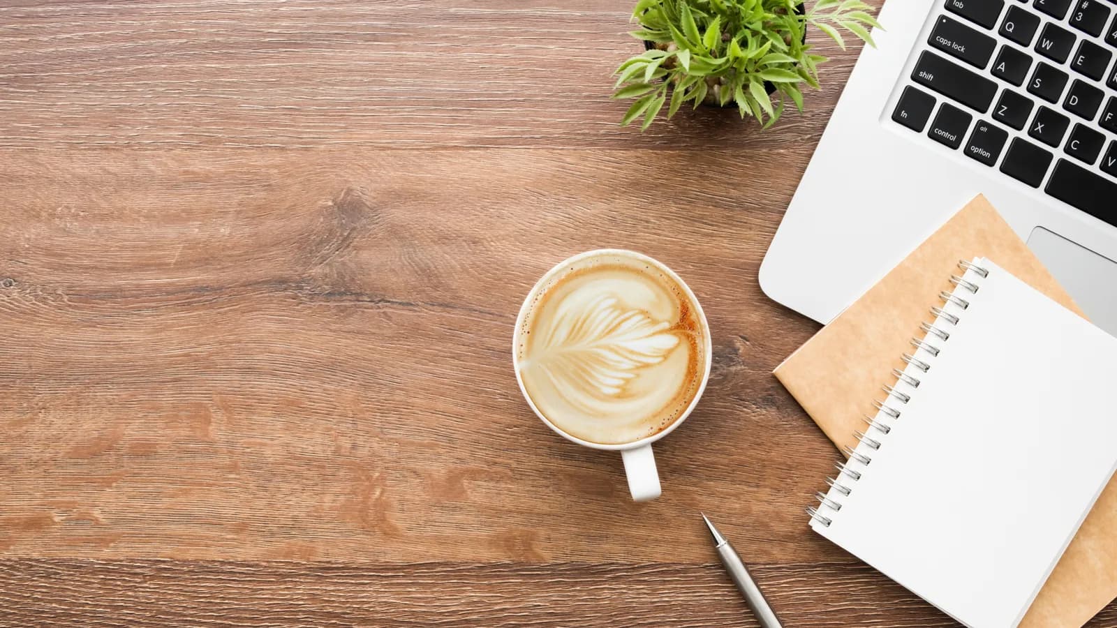 A cup of coffee with latte art beside a laptop, a notebook and a pen on a wooden desk