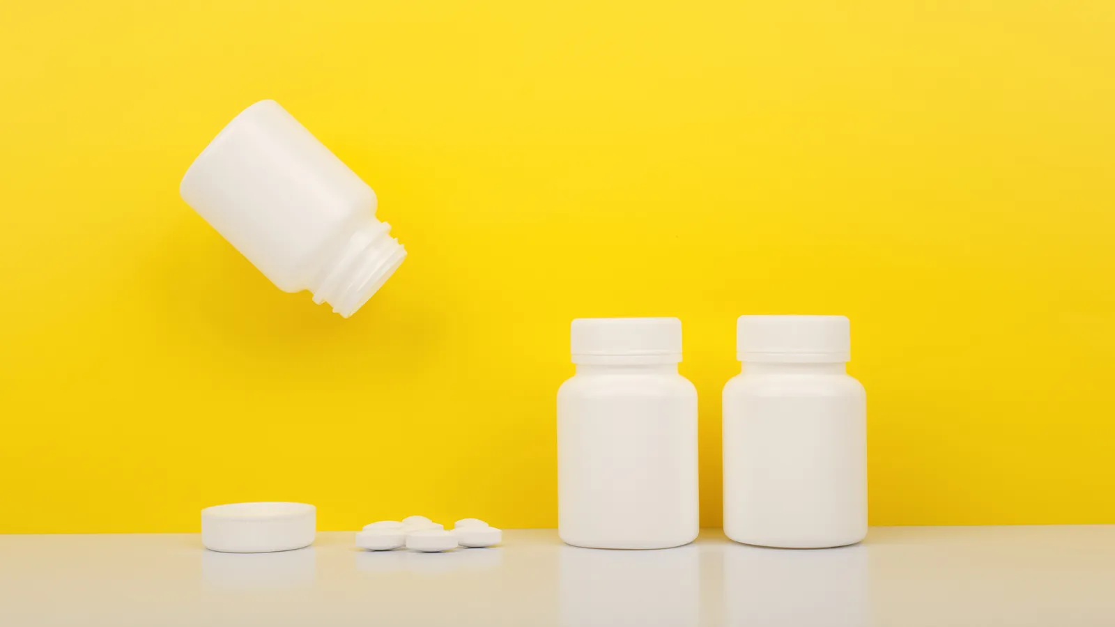 Three white medicine bottles on a white table against a yellow background, one on its side with a few tablets beside it