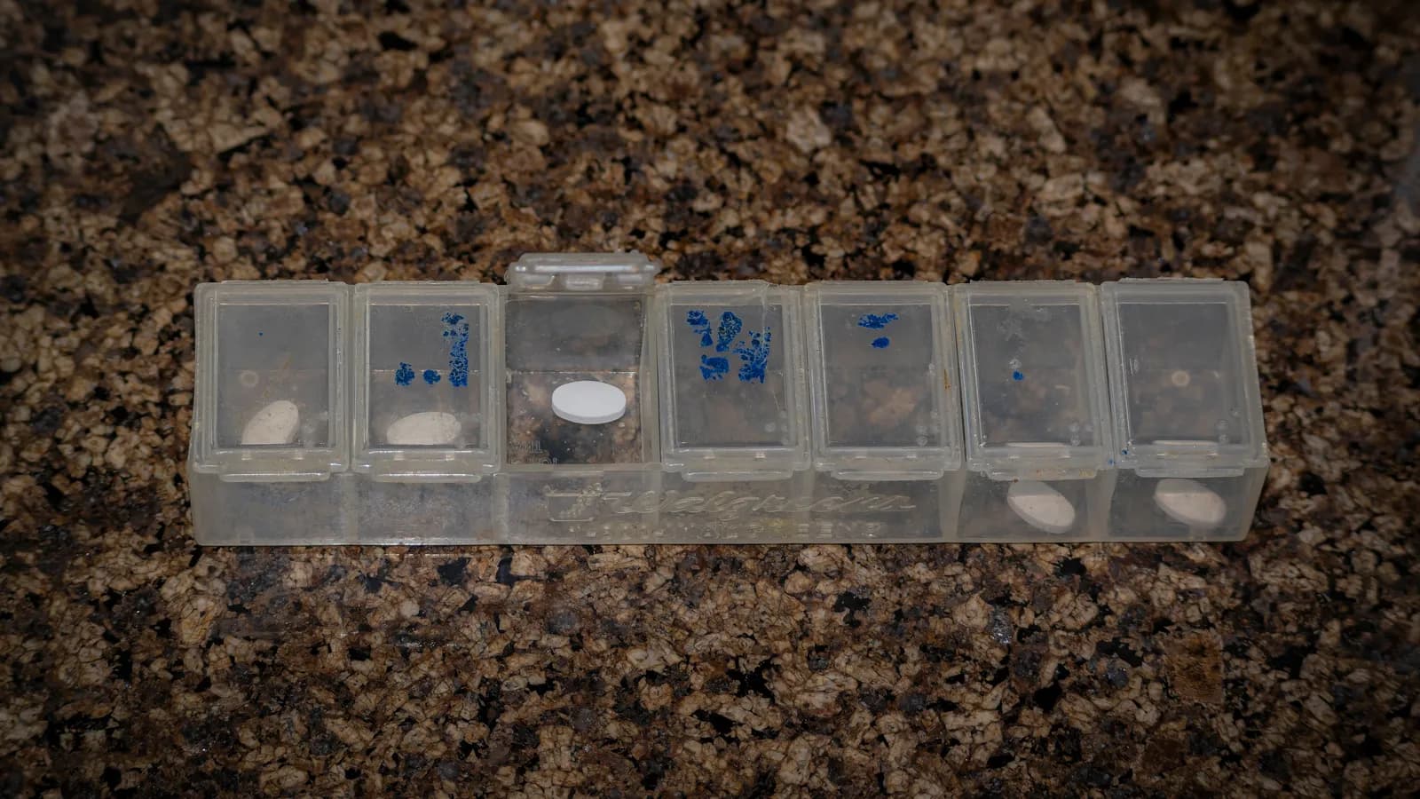 A clear weekly pill organizer with its lids open and tablets in several compartments on a dark granite counter