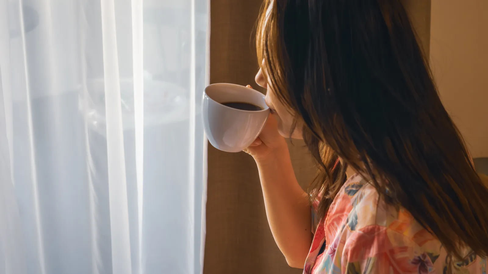 A woman seen from the side holding a cup of coffee and looking out through a sheer curtain