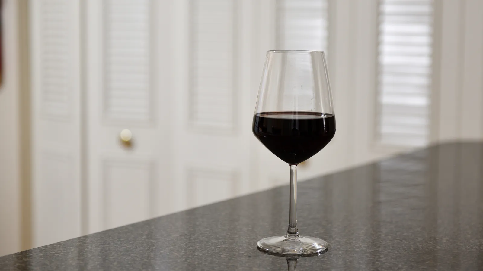 A single glass of red wine on a dark counter in front of white shutters